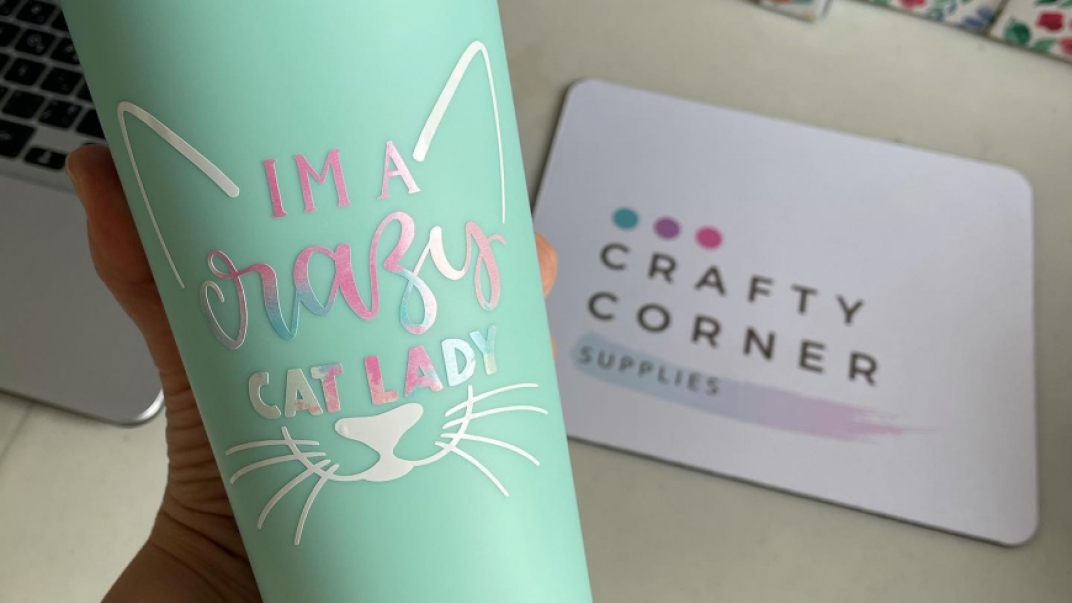 Metal mug with a cat graphic and the words "I am a crazy cat aldy" written on it. In the background there is a mat with the words" Crafty Corner" on it.