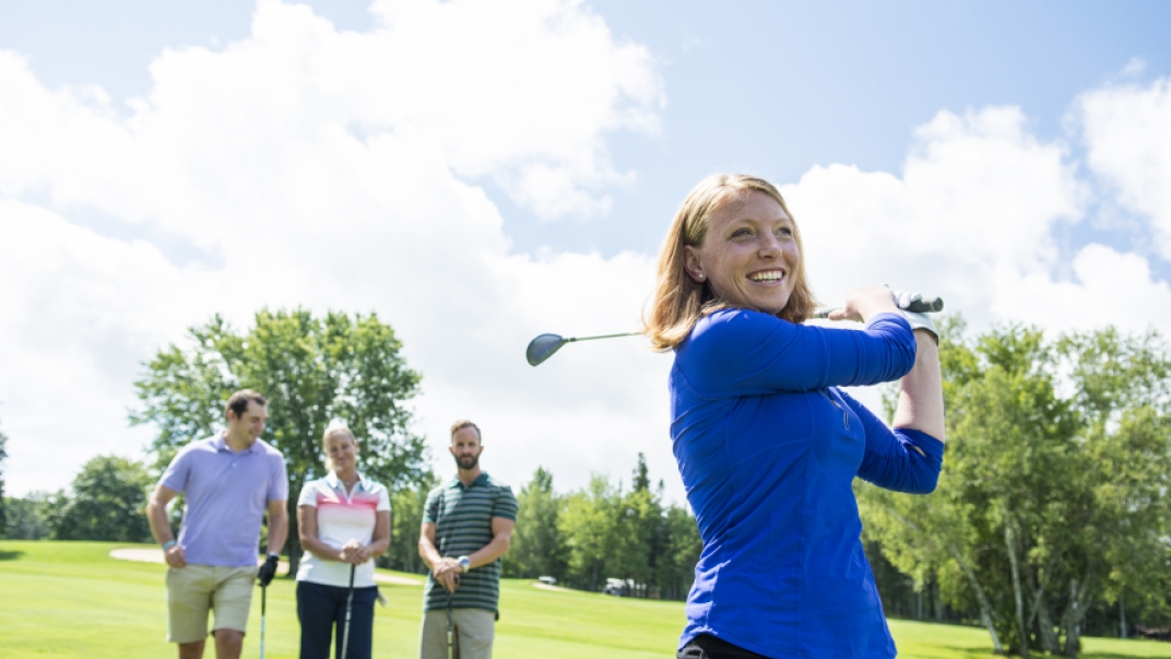 white woman in bright blue long sleeve shirt swings a golf club while her companions watch in the background