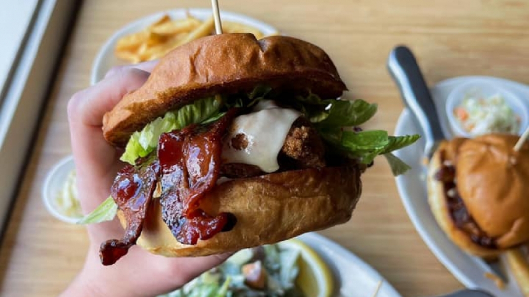 Someone holding a bacon cheeseburger close to a camera with burgers and fries on the table in the background