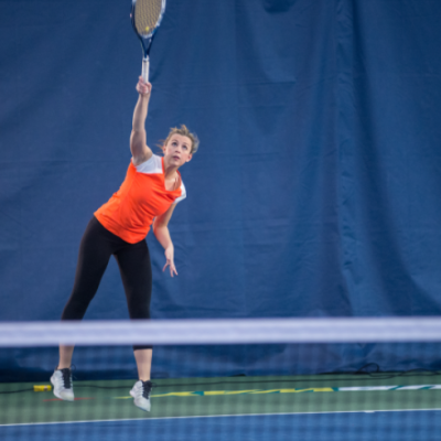 Woman in orange shirt playing tennis at a tennis court.