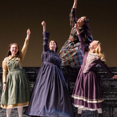 four women in period clothing raise their fist on a theatre stage