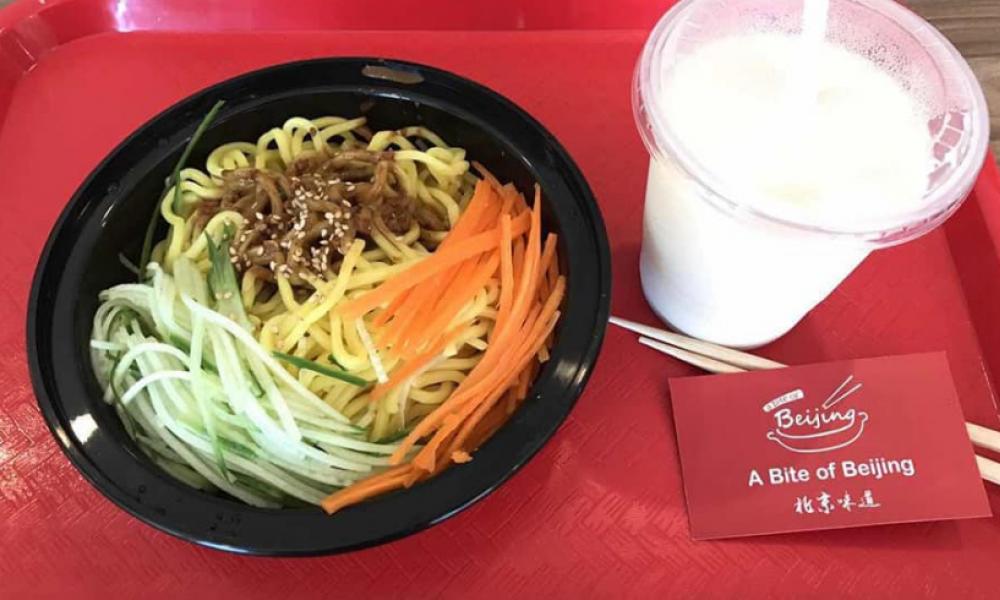 A red cafeteria tray with a black take out container of noodles and julienned cucumbers and carrots on it. Also on the tray is a to go cup holding a white liquid, chopsticks and a business card.