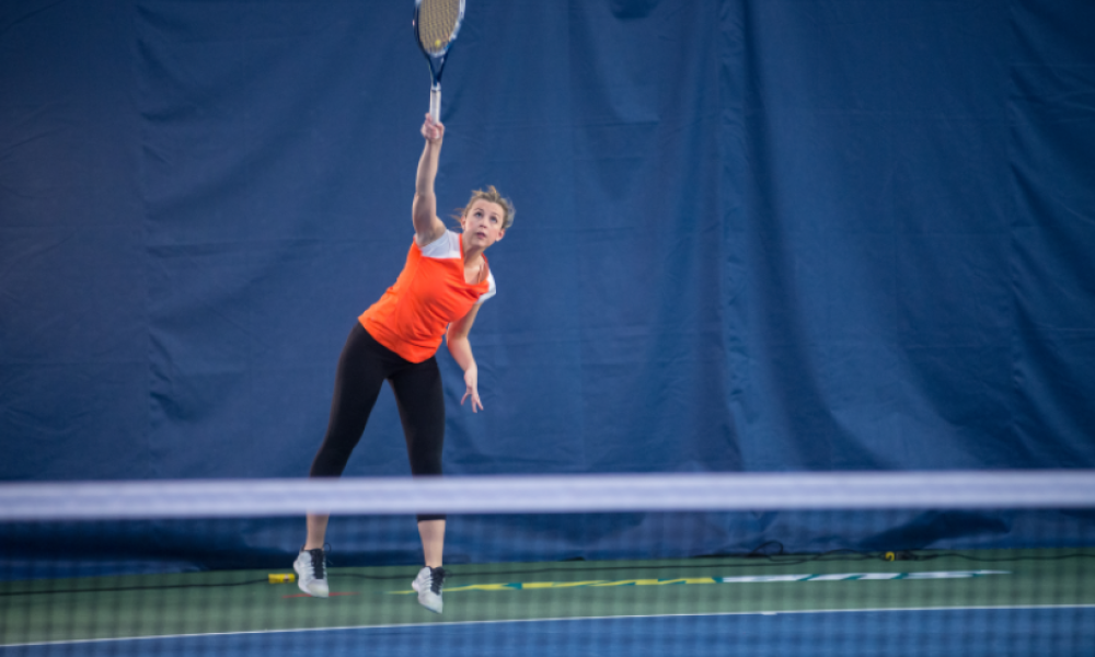 Woman in orange shirt playing tennis at a tennis court.