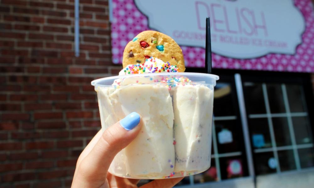 A hand holding a cup of vanilla ice-cream with a cookie on top in front of the Delish store sign. 