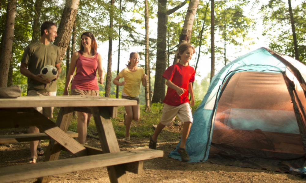 Smiling family playing at a campsite in a forest with a tent and picnic table. 