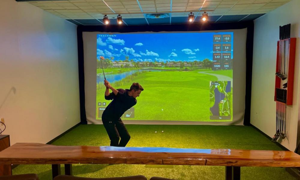 Man swinging golf club at golf ball. There is an electronic golfing projection on a screen in the background