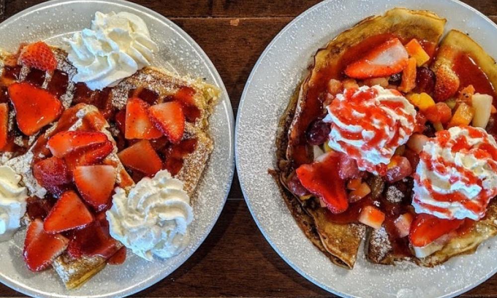 A plate of waffles and a plate of crepes both with strawberries, whipped cream and icing sugar on them